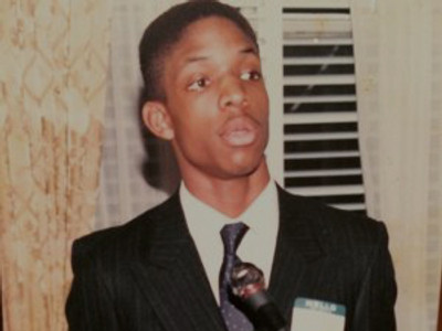 A young Jay Cameron in a suit speaking into a microphone
