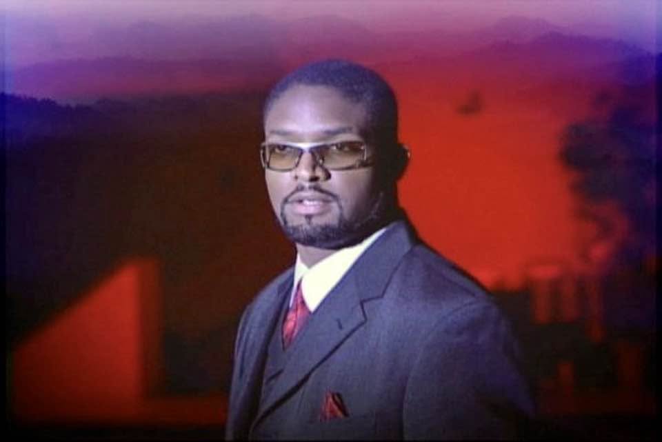 Jay Cameron in a dark suit and tinted glasses in front of a red and purple backdrop