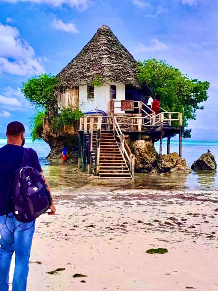 Jay Cameron walking across the sand toward a thatched restaurant built on a rock in the sea