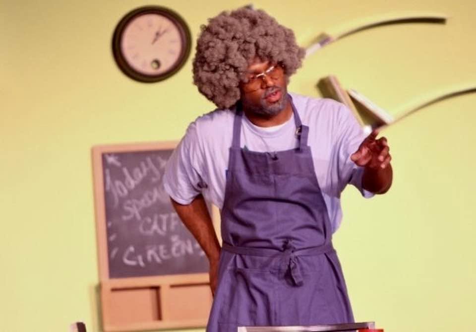 An actor in an apron and wig working in a kitchen set during a live production