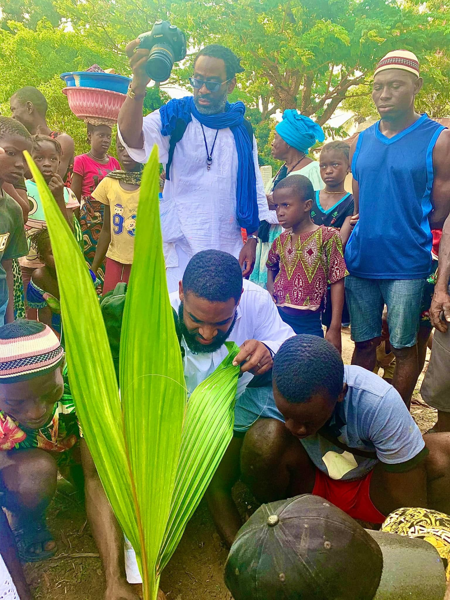 Jay Cameron planting a palm seedling with a village gathered around in Sierra Leone