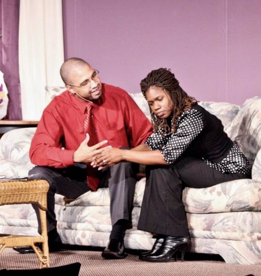 Two actors seated on a sofa in a tense scene on a living room set