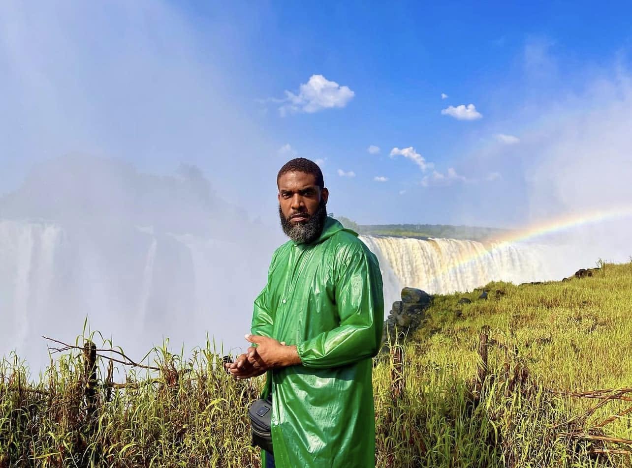 Jay Cameron in a green rain jacket at Victoria Falls with a rainbow over the water
