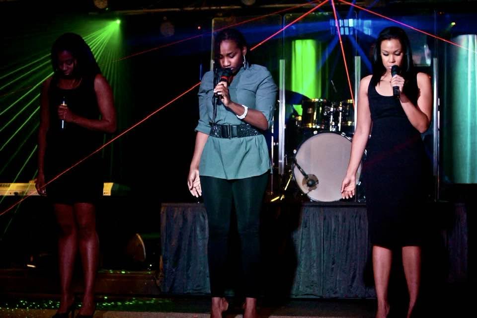 Three performers with microphones on a stage lit by green and red lasers